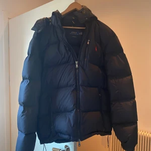 Mörkblå dunjacka Polo Ralph Lauren XL - Säljer en mörkblå dunjacka från Polo Ralph Lauren i storlek XL. Jackan har en klassisk pufferstil med huva, dragkedja framtill och flera fickor. Ikonisk röd logga på bröstet och snygg, sportig look. Perfekt för kalla vinterdagar. Passar för dig som är runt 190 cm lång. Skriv för frågor och pris