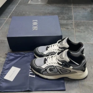 Dior sneakers i svart, grå och vit - Snygga Dior sneakers med coola detaljer i svart, grått och vitt. Skorna har tydliga CD-loggor på sidan och hälen, meshpaneler och skinnpartier. Klassisk snörning och chunky sula ger en modern vibe. Perfekta för dig som vill sticka ut med exklusiva sneakers.