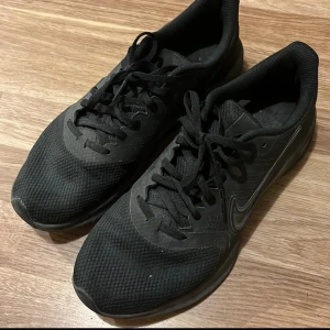 Svarta Nike sneakers strl 43 - Svarta Nike sneakers med mesh-ovandel och klassisk swoosh på sidan. Skorna har rund tå, platt sula och snörning. Perfekta för dig som gillar sportig stil och vill ha bekväma skor till träning eller vardags. Storlek EU 43, US 9.5. Pris kan diskuteras.