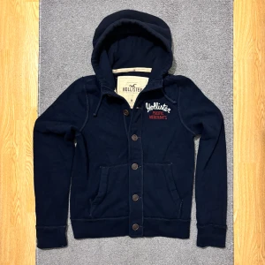 Blå Vintage Hollister Kofta/Cardigan Med Knappar - Mörkblå hoodie från Hollister med broderad logga på bröstet i vitt och rött. Tröjan har stora knappar framtill, huva med snörning och två fickor. Perfekt för dig med avslappnad stil, perfekt för kyligare dagar. Modell 174-175. Hör av dig vid ytterligare frågor!