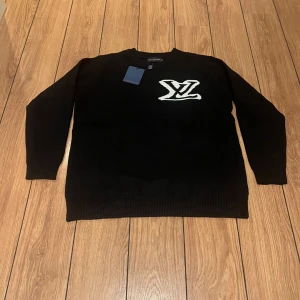 Svart stickad tröja Louis Vuitton - Svart stickad tröja från Louis Vuitton med stort vitt LV-logo på bröstet. Tröjan har rund halsringning, ribbade muddar och är långärmad. Perfekt för dig som vill ha en clean och lyxig look med streetvibe. Priset kan sänkas för att den är andvänd