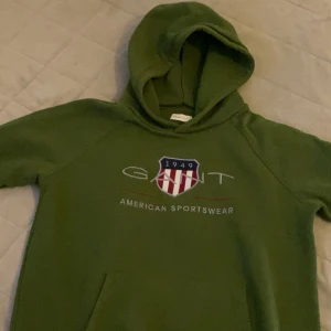 Grön hoodie från GANT med tryck - Säljer en grön hoodie från GANT med klassiskt tryck och emblem framtill. Tröjan har huva och känguruficka, perfekt för chill dagar. Mjuk och skön bomullskänsla, passar dig som gillar sportig och avslappnad stil.