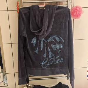 Vintage Juicy Couture mörkblå velour hoodie - Juicy Couture hoodie i mörkblå velour med glittrande stenar och stor logga på ryggen. Klassisk huva, dragkedja framtill med J-detalj och kängurufickor. Perfekt för dig som älskar 00-talsvibbar och vill ha en mjuk och snygg tröja.