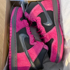 nike sb dunk high run the jewels - Nike SB Dunk High Run The Jewels är en exklusiv och kraftfull sneaker med en ovandel i mörkrosa och lila mocka, kombinerad med en metallic Swoosh som ger ett unikt uttryck. Modellen har rund tå, platt sula och klassisk snörning, och levereras med fyra olika skosnören i rosa, gult, grått och grönt, så du kan variera looken efter smak. Dessutom kan du byta ut de metallfärgade snörbrickorna från guld till svart för en mer personlig stil.  Skorna är helt nya och aldrig använda.