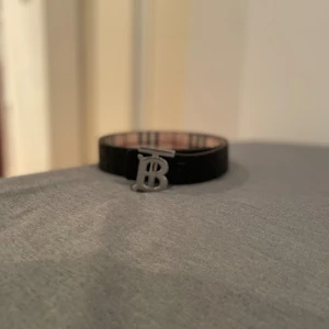 Burberry bälte med rutigt mönster - Säljer ett snyggt Burberry bälte med klassiskt rutigt mönster i beige, svart och rött. Bältet har en silverfärgad metallspänne med Burberrys ikoniska 'B'-logga. Perfekt accessoar för att lyfta din outfit med lite lyxig vibe.