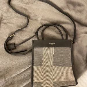 David Jones axelväska / handväska med mönster - Snygg axelväska från David Jones i mörkbrun och beige med grafiskt rutigt mönster. Väskan har både handtag och justerbar axelrem, samt innerfack med dragkedja. Väskan är aldrig använd. 