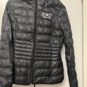 Svart dunjacka från EA7 Emporio Armani - Snygg svart dunjacka från EA7 Emporio Armani med huva och dragkedja framtill. Jackan har quiltad design och logga på bröstet. Perfekt för kyliga dagar och ger en clean, sportig look.