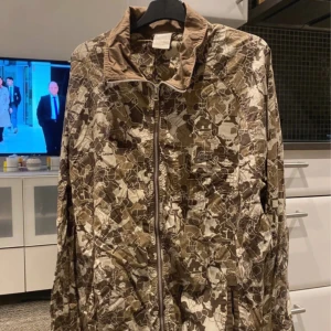 Brun camo vindjacka med dragkedja - Säljer en brun och beige vindjacka med camouflagemönster. Jackan har hel dragkedja framtill, hög krage och två fickor med dragkedja. Lätt material och elastiska muddar vid ärmslut och nederkant. Perfekt för dig som gillar streetstyle och vill sticka ut.