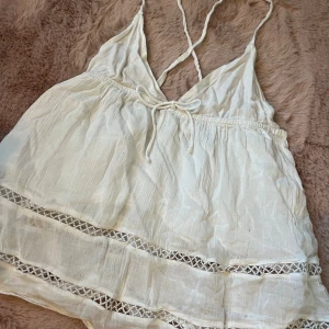 Vit linnetopp med spets från Gina Tricot - Supersöt vit linnetopp från Gina Tricot med tunna axelband som korsas i ryggen och knyt fram. Toppen har fina spetsdetaljer och är gjord i ett lätt, luftigt material. Perfekt för varma dagar och ger en boho-vibe till din outfit. Storlek 158-164!