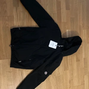 Svart vindjacka från Moncler med huva - Svart vindjacka från Moncler med huva, hel dragkedja och två fickor med dragkedja framtill. Jackan har Moncler-logga på ärmen och är tillverkad i ett lätt, syntetiskt material. Perfekt för dig som vill ha en stilren och sportig look.