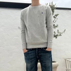 Grå stickad tröja (HELT NY) - (Modellen är 183)Snygg grå stickad tröja från Polo Ralph Lauren med klassisk rund hals och ribbade muddar. Tröjan har ett mörkblått broderat Polo-logo på bröstet och är perfekt för dig som gillar stilrena och enkla plagg. Passar dig som vill ha en normal passform och långärmad modell.