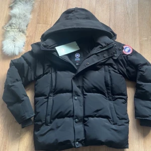 Svart dunjacka från Canada Goose S - Säljer en svart dunjacka från Canada Goose med avtagbar huva och pälskant. Jackan har klassisk patch på ärmen, dragkedja och knappar framtill samt två stora fickor. Perfekt för kalla vinterdagar och riktigt snygg streetstil. OBS! En knapp är sönder men jag har kvar knappen så jag tror det går att lagra