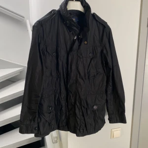 Svart vindjacka från Polo Ralph Lauren - Svart vindjacka från Polo Ralph Lauren i storlek M. Jackan har flera fickor med knappar, hög krage och axelklaffar. Tillverkad i glansig polyester med dragkedja och knappstängning framtill. Perfekt för dig som gillar stilren och funktionell design.