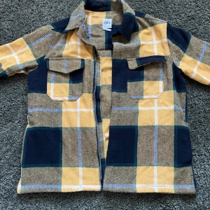Rutig overshirt från Zara i gult och blått - Overshirt från Zara i relaxed fit med stort rutmönster i gult, blått och beige. Jackan har två bröstfickor med lock, klassisk krage och dragkedja framtill. Perfekt lager-på-lager-plagg med lång ärm och mjukt material.