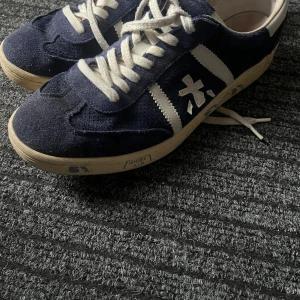 Premiata blå sneakers med mocka - Snygga sneakers från Premiata i mörkblå mocka med vita detaljer och snörning. Skorna har en klassisk låg profil, rund tå och vit sula med branding på sidan. Perfekt för dig som gillar retrostil och vill sticka ut med ett coolt märke. OBS! Ordinarie pris är 3000 kr