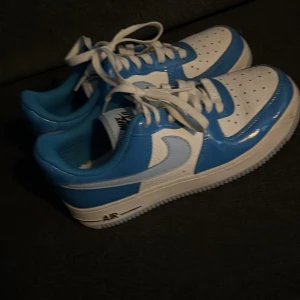 Nike Air Force 1 blå och vita sneakers - Säljer ett par Nike Air Force 1 sneakers i blått och vitt med klassisk låg siluett. Skorna har snörning, perforerad tå och platt sula. Ovandelen är i skinn med blanka blå paneler och vit bas, samt ljusblå swoosh på sidorna. Perfekt för dig som gillar streetwear och snygga färgkombinationer.