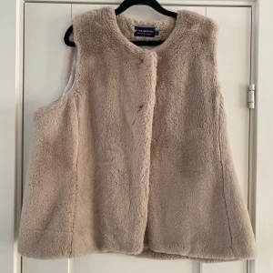 Beige pälsväst från River Woods XL - Fluffig och mjuk beige pälsväst från River Woods i storlek XL. Västen har en rak och loose passform, rund halsringning och diskret knäppning framtill. Insidan är slät och ljus, vilket gör den extra skön att bära. Perfekt för att addera lite extra värme och stil till din outfit. Mått Över bröstet: ca 59 cm Längd från axeln och ner: ca 68 cm  100% polyester  Kommer från ett djur och rökfritt hem.