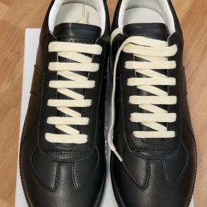 Maison Margiela svarta sneakers strl 43 - Maison Margiela sneakers i svart skinn med vit snörning och rund tå. Klassisk och stilren design med detaljerad sula och diskret branding under foten. Kommer med originalpåse från Maison Margiela Paris. Perfekt för dig som gillar exklusiva och minimalistiska sneakers.