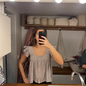 Virala Beige blus med volangärmar från Zara - Supersöt beige blus från Zara med breda axelband och volangdetaljer på ärmarna. Blusen har knappar framtill och en luftig, flowy passform. Tillverkad i ett tunt, strukturerat bomullstyg som känns lätt och somrigt. Perfekt för dig som gillar romantiska vibes!