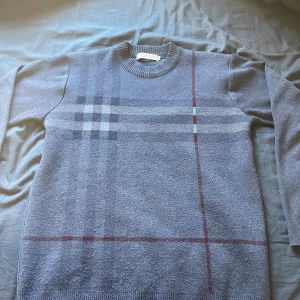 Blå rutig stickad tröja Burberry - Snygg blå stickad tröja från Burberry med klassiskt rutigt mönster i grått och rött på framsidan. Rund halsringning och långa ärmar med ribbade muddar. Perfekt för dig som gillar stilrena och tidlösa plagg med premiumkänsla.