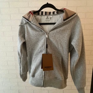 Grå Burberry Hoodie med dragkedja - Snygg grå hoodie från Burberry med hel dragkedja och klassiskt rutigt foder i huvan. Tröjan har snörning i huvan, ribbade muddar och känguruficka framtill. Perfekt för dig som vill ha en stilren och lyxig vardagslook. Olika storlekar, pris kan oxå diskuteras!;/