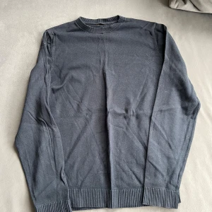 Mörkgblå stickad tröja med rund hals - En stilren mörkgrå stickad tröja med rundad hals och ribbade muddar vid ärmslut och nederkant. Perfekt för dig som gillar enkla och klassiska plagg. Långärmad och mjuk i materialet, passar till många olika outfits.