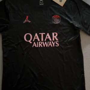 Psg fotbollströja - Snygg svart PSG fotbollströja från Jordan med rosa detaljer och Qatar Airways-tryck på bröstet. Kortärmad modell i lätt, tekniskt material som andas. PSG-logga och Jumpman-logga på framsidan. Perfekt för dig som vill ha en stilren och modern look på planen.