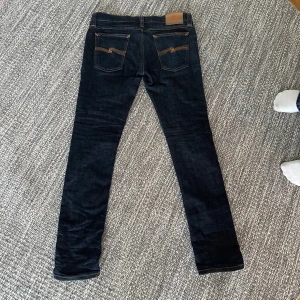 Mörkblå jeans från Nudie Jeans, W32 L32 - Snygga mörkblå jeans från Nudie Jeans med klassisk orange söm och logga på bakfickan. Modellen har smal passform och raka ben, tillverkade i slitstarkt jeanstyg. Perfekta för dig som gillar stilrena och tidlösa jeans.