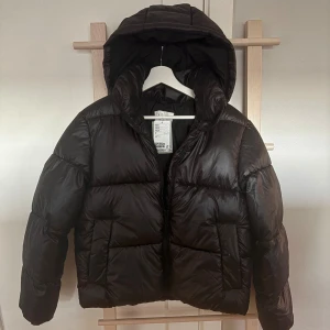 Svart pufferjacka från H&M med huva - Svart pufferjacka från H&M i storlek S. Jackan har en oversized look med huva och dragkedja framtill. Helt oanvänd med lappen kvar,          nypris 449kr