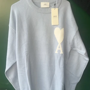 Ljusblå stickad tröja från Ami - Superfin ljusblå stickad tröja från Ami med stort vitt hjärta och bokstaven A på bröstet. Tröjan har rund halsringning, ribbade muddar och en relaxed passform. Perfekt för dig som gillar stilrena och trendiga plagg med en twist.
