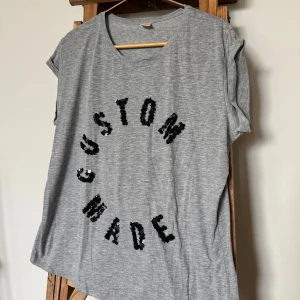 Grå t-shirt med paljetter från Custommade - Gråmelerad t-shirt från Custommade med svart paljetttext 'CUSTOM MADE' på framsidan.