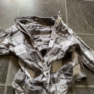 Gråvit rutig skjorta med knappar - Säljer en grå och vit rutig skjorta med långa ärmar och knappar framtill. Skjortan har klassisk krage och bröstficka, tillverkad i mjukt bomullstyg. Perfekt för en avslappnad och stilren look.