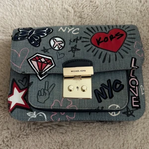 Michael Kors handväska med graffiti - Cool handväska från Michael Kors i denim-look med graffitiinspirerade tryck som hjärtan, fjäril, diamant och text. Väskan har guldfärgat lås framtill och flera fack inuti. Färgerna går i grått, rött, vitt, svart och rosa. Perfekt för dig som vill sticka ut med din stil.