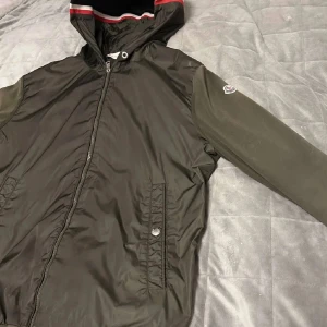 Grön vindjacka från Moncler med huva - Snygg grön vindjacka från Moncler med huva och dragkedja framtill. Jackan har Moncler-logga på ärmen, ribbade muddar och detaljer i rött och vitt på insidan av huvan. Materialmix med polyester och stickade ärmar för en sportig vibe.