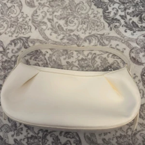 Vit axelväska  - Stilren vit axelväska i syntetmaterial med mjukt rundad form och silverfärgad dragkedja upptill. Väskan har ett minimalistiskt uttryck och passar perfekt för dig som gillar enkel och clean design. Rymmer det viktigaste och är lätt att bära över axeln.