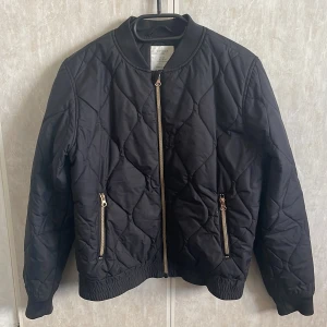 Svart quiltad bomberjacka från Lindex - Svart quiltad bomberjacka från Lindex med guldfärgade dragkedjor och ribbade muddar. Jackan är vattenavvisande och har två fickor framtill med dragkedja. Perfekt för dig som vill ha en stilren och bekväm jacka med snygg passform.