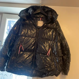 Svart pufferjacka från Tommy Hilfiger - Säljer en glansig svart pufferjacka från Tommy Hilfiger med luva och dragkedja. Jackan har två fickor med snygga röda och vita detaljer vid dragkedjorna. Perfekt för kalla dagar och har en riktigt modern vibe.