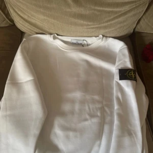 Vit sweatshirt från Stone Island - Vit sweatshirt från Stone Island med klassisk rund hals och ikonisk logga på vänster ärm. Tröjan är i mjuk bomull och har en clean look som passar perfekt till jeans eller trackpants. Enkel och stilren design med långa ärmar.