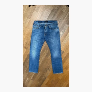 Nudie Jeans Average Joe - Snygga blå jeans från Nudie Jeans med klassisk femficksdesign och orange sömmar. Jeansen har raka ben med råa, avklippta benslut och en broderad bakficka. Perfekta för en avslappnad och trendig look.