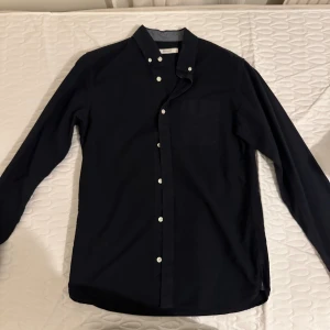 Mörkblå skjorta från Premium - Snygg mörkblå skjorta från Premium med klassisk button down-krage och vita knappar. Skjortan har en bröstficka och är långärmad, perfekt för en stilren look. Tillverkad i mjuk bomull som känns skön mot huden.