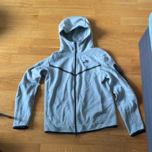 Grå Tracksiut med huva från Nike - Nike Tracksiut i ljusgrått med svart logga på bröstet och svarta detaljer. Jackan har hel dragkedja, huva och en extra ficka med dragkedja på ärmen. Tillverkad i mjukt syntetmaterial, perfekt för chill eller träning.pris kan diskuteras