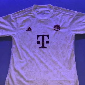 Vit blommig bayern fotbollströja Adidas L - Snygg vit fotbollströja från Adidas i storlek L med blommigt mönster och rödbruna detaljer. Tröjan har korta ärmar, rund hals och är tillverkad i lätt, ventilerande Aeroready-material. Tryck med T-Mobile-logga och klubbmärke på bröstet.