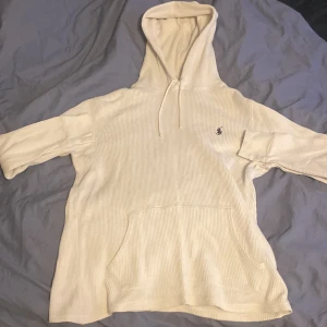 Vit hoodie från Polo Ralph Lauren - Vit hoodie från Polo Ralph Lauren med klassisk broderad logga på bröstet. Tröjan har huva med snörning, känguruficka och är tillverkad i ribbad bomull. Perfekt för chill dagar och snygg till jeans eller joggers. 