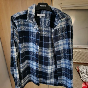 Blå och vit rutig skjorta från SMOG - Snygg rutig skjorta från SMOG i blå och vit ton. Skjortan har klassisk krage, långa ärmar och knappar framtill. Materialet är mjukt och lite tjockare, perfekt för lager-på-lager. Passar dig som gillar en chill och avslappnad stil.