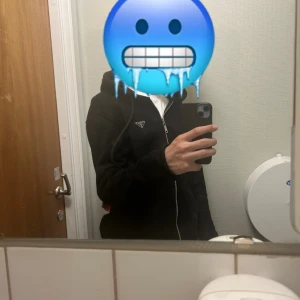 Svart hoodiejacka från Prada - Svart hoodiejacka från Prada med dragkedja framtill och justerbar huva. Jackan har en klassisk passform och är tillverkad i mjukt material, perfekt för en avslappnad och stilren look. Diskret Prada-logo på bröstet.