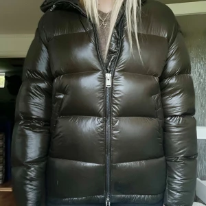 Grön pufferjacka med huva - Säljer en grön pufferjacka med glansig finish och huva. Jackan har en rejäl dragkedja framtill och breda, vadderade paneler som ger en riktigt fet look. Perfekt för kalla dagar när du vill hålla värmen och ändå se snygg ut. Jättebra skick och slutsåld 
