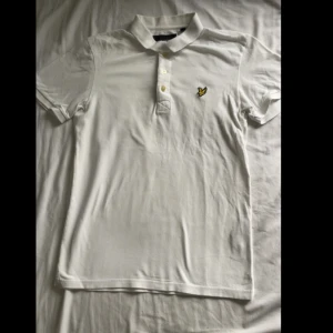Vit piké från Lyle & Scott - Vit pikétröja från Lyle & Scott med klassisk krage, tre knappar och den ikoniska gula örnloggan på bröstet. Tillverkad i mjuk bomull, perfekt för en clean och stilren look.