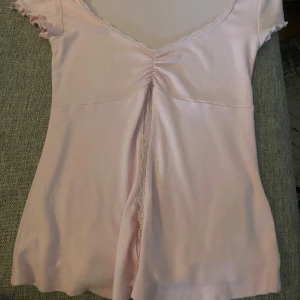 Ljusrosa topp med spets från Gina Tricot - Supersöt ljusrosa topp från Gina Tricot i storlek XS. Toppen har v-ringning med spetskant, rynkdetalj framtill och korta ärmar med volangkant. Lite nopprig på ryggen.
