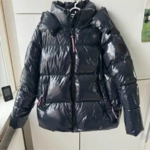 Jacka - Snygg svart pufferjacka från Tommy Hilfiger i storlek L. Jackan har glansig finish, huva och dragkedja framtill. Klassisk quiltad look och två fickor med dragkedja. Perfekt för kalla dagar. Köpte på vinted men är för stor