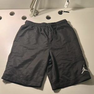 Svarta Jordan shorts med logga - Säljer ett par svarta Jordan shorts med vit broderad Jumpman-logga på benet. De har elastisk midja med snörning och är gjorda i lätt polyester med meshfoder för extra komfort. Perfekta för sport eller chill. Snygg och enkel design som alltid funkar.